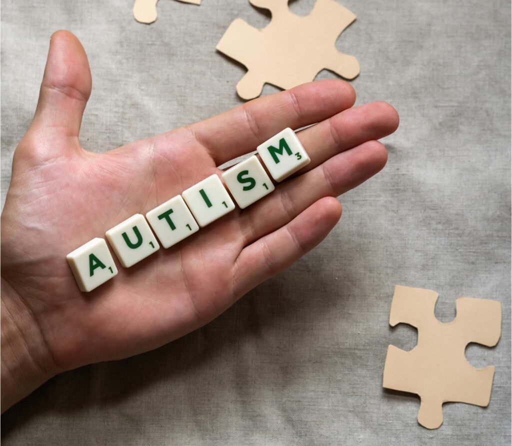 A hand holding lettered tiles that spell AUTISM, with jigsaw puzzle pieces nearby