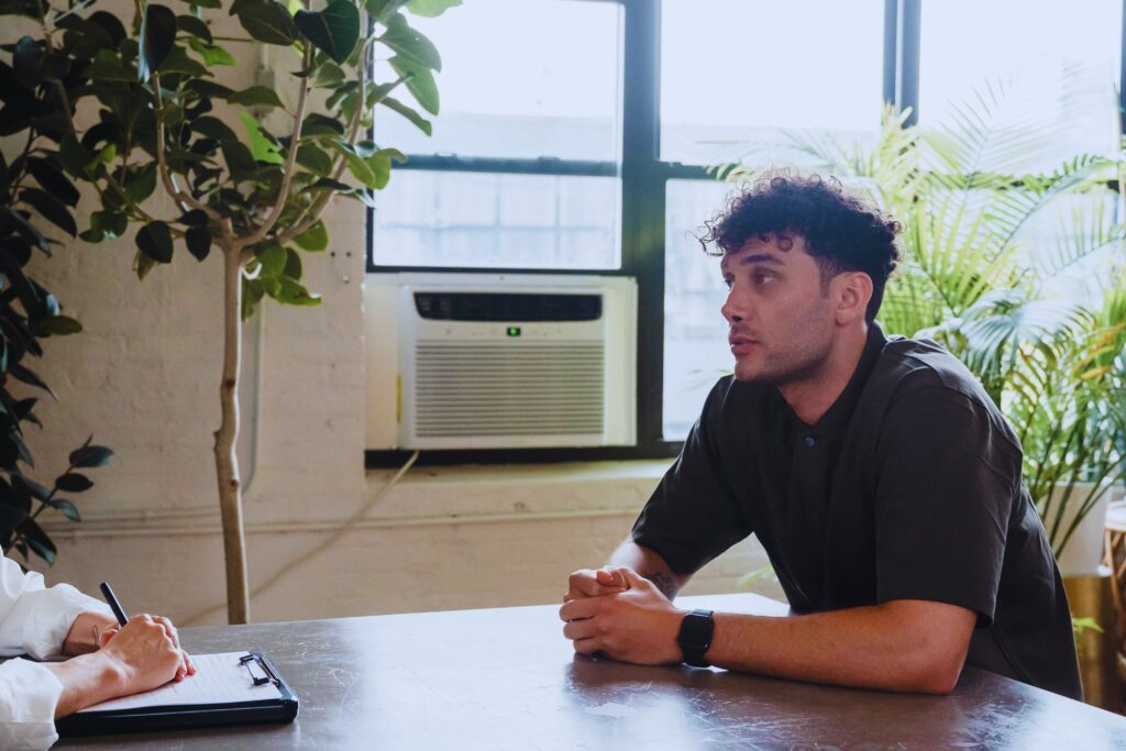 A young man talking with a clinician who takes notes during a session in a plant-filled office